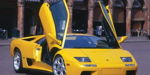 Żółty Lamborghini Diablo z otwartymi drzwiami, zaparkowany na brukowanej ulicy przed budynkami z arkadami.