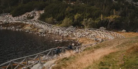 Dwie osoby idą ścieżką przy jeziorze, w kierunku schroniska Samotnia. Drewniane balustrady prowadzą wzdłuż kamienistego brzegu.