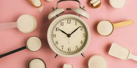 Wecker umgeben von Löffeln mit Kollagenpulver und Kapseln. Wann ist die beste Zeit, kollagen morgens oder abends einzunehmen?