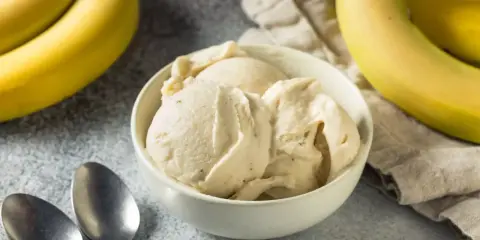 Un cremoso helado de plátano servido en un cuenco blanco, con plátanos amarillos frescos al fondo.