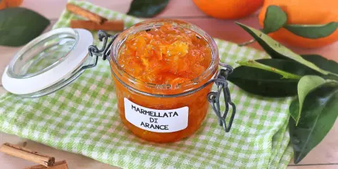 Barattolo di marmellata di arance fatto in casa, con arance fresche e bastoncini di cannella sullo sfondo.