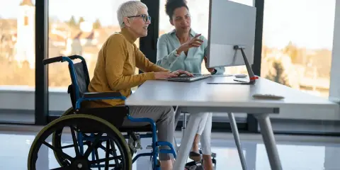 Kobieta na wózku inwalidzkim i jej koleżanka pracują przy komputerze, pokazując, że niepełnosprawność sprzężona nie jest przeszkodą w pracy.