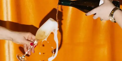 Schaumiges Sektglas läuft über, während eine Flasche auf orangefarbenem Hintergrund eingegossen wird. Eine Szene, die an die **zero dosage definition** erinnert, wo jeder Tropfen zählt.