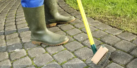 Person en bottes vertes brossant la mousse sur une terrasse pavée. Astuce pour savoir comment enlever la mousse sur une terrasse.