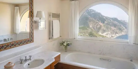 Un bagno di lusso con vasca, lavandino e specchio decorato, che offre una vista mozzafiato sulle montagne e sul mare. Tra i bagni più belli ed eleganti.