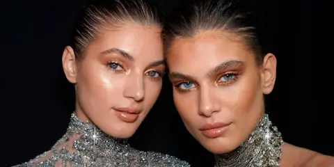Dos mujeres con maquillaje dorado y un sutil rabillo del ojo, luciendo collares brillantes.