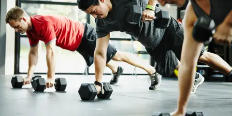 Drei Personen machen eine Plank-Übung mit Kurzhanteln. Ein Mann in Rot, ein Mann mit Kappe und eine Frau in Grau trainieren. CrossFit gesund und stark.