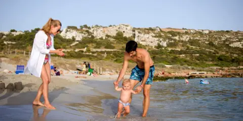 Familia disfrutando de un día en la playa en Menorca con niños. El padre ayuda a su bebé a dar sus primeros pasos en el agua, mientras la madre observa sonriente.