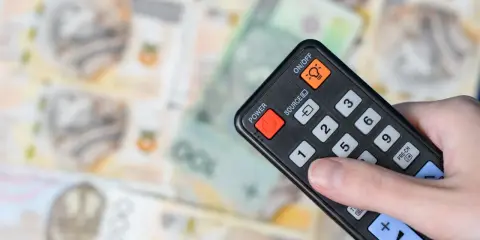 Dłoń trzyma pilota na tle polskich banknotów. Czy abonament RTV i zameldowanie mają związek z kosztami?