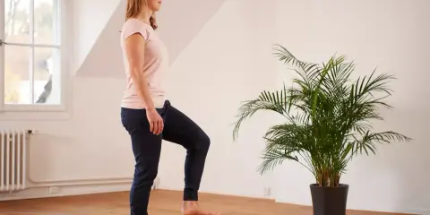 Frau übt auf einem Bein stehen im hellen Raum, neben einer grünen Palme.