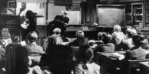 Lehrer mit Stock ermahnt Schüler in einer Klasse, die an eine Schule im Mittelalter erinnert.