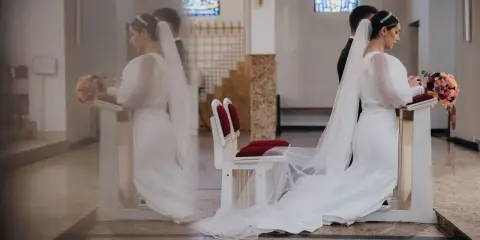 Para młoda klęczy przy ołtarzu. W tle witraże i ławki. Czyżby to były przesądy ślubne dla pana młodego?