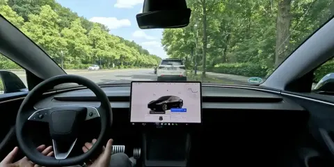 Interno di una Tesla in movimento in Europa, con il sistema FSD attivo. La strada è fiancheggiata da alberi verdi.