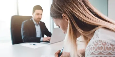 Kobieta podpisuje dokumenty, planując jak zmienić nazwisko po rozwodzie. Mężczyzna w tle pracuje przy laptopie.