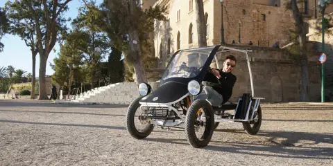 Ein Mann fährt einen Duo Moke durch eine sonnige Gasse mit historischen Gebäuden im Hintergrund.