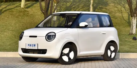 Auto elettrica piccola 4 posti, bianca, con tetto nero, parcheggiata su un vialetto pavimentato con alberi sullo sfondo.