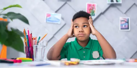 Niño frustrado con las manos en la cabeza, rodeado de materiales de arte, quizás lidiando con problemas de aprendizaje.
