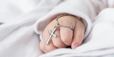 Dziecięca dłoń trzyma krzyżyk na łańcuszku, symbol tego, co jest potrzebne do chrztu.