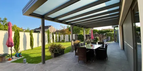 Une belle pergola moderne avec un toit vitré pour couvrir l'espace repas extérieur. Le jardin est bien entretenu, avec une pelouse verte et des cyprès élancés.