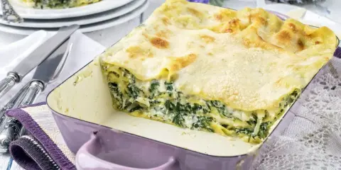 Pyszne lasagne ze szpinakiem w fioletowym naczyniu, gotowe do podania. Obok talerze z porcją i sztućce.