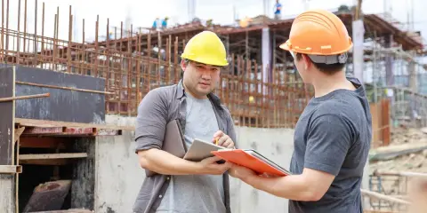 Dwóch budowlańców w kaskach omawia plany na placu budowy. Jeden z nich trzyma tablet, drugi teczkę z dokumentami.