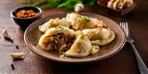 Talerz pełen pysznych pierogów z kapustą i grzybami, posypanych szczypiorkiem. Obok czosnek i pieprz.