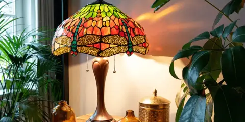 Lampe tiffany origine, avec abat-jour en vitrail coloré orné de libellules, posée sur une table en bois près de plantes vertes.
