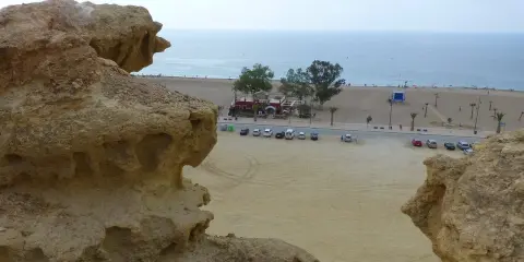 Rocas erosionadas en primer plano, con la playa de Bolnuevo, el mar y un paseo marítimo con palmeras al fondo.