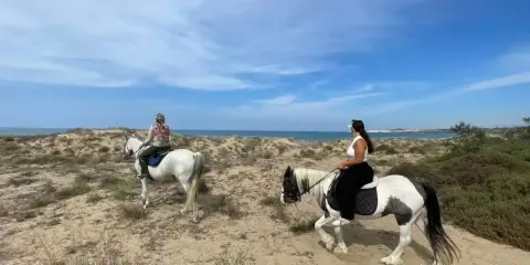 Due donne godono di una passeggiata a cavallo in Sicilia, tra dune sabbiose e il mare azzurro.