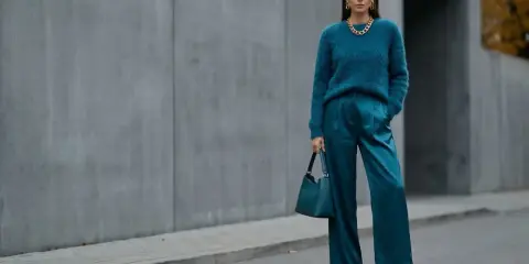 Kobieta w eleganckim, futrzanym swetrze i satynowych spodniach w kolorze petrol. Torebka i buty również w tym samym odcieniu.