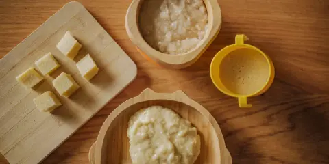 Pierwsza kaszka dla niemowlaka karmionego piersią. Na drewnianym blacie leżą kostki banana, miseczka z kaszką i żółty kubeczek.