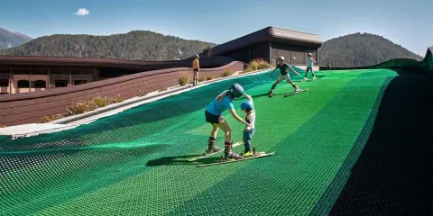 Bambini imparano a sciare su una pista verde al Falkensteiner Family Resort Lido, con montagne sullo sfondo.