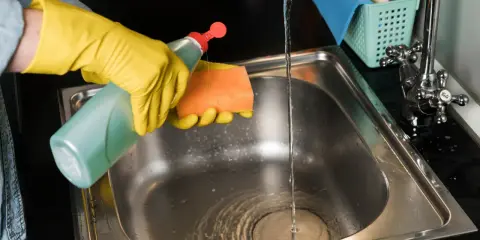 Rękawiczki i płyn do naczyń pomogą Ci dowiedzieć się, czym wyczyścić zlew ze stali nierdzewnej. Woda z kranu spływa do czystego zlewu.