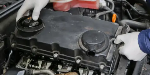 Mechanik w białych rękawiczkach sprawdza silnik, odkręcając korek wlewu oleju. Widoczne elementy silnika i układu odmy.