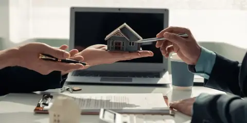 Deux mains présentent une maquette de maison, symbolisant comment devenir investisseur immobilier. Un contrat et un ordinateur sont visibles.