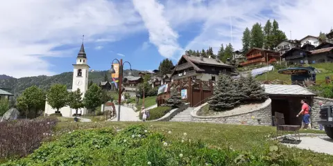 A picturesque village in Chamois, Italy, with a church tower, wooden chalets, and lush greenery. Scopri cosa vedere in questo incantevole borgo alpino.