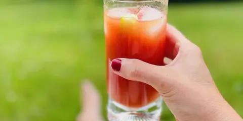A hand with red nail polish holds a tall glass of Bloody Mary with ice and a cucumber slice, outdoors.