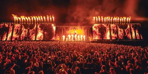 Un'immensa folla partecipa a un festival tekno, illuminata da fiammate spettacolari e schermi con immagini di fuoco.