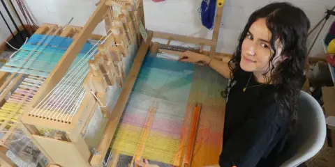 Une jeune femme souriante, passionnée par son tissier métier, travaille sur un métier à tisser en bois. Les fils colorés créent une œuvre vibrante.