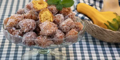 Un tazón lleno de buñuelos de calabaza cubiertos de azúcar, con una ramita de menta encima. Al fondo, calabazas en una cesta.