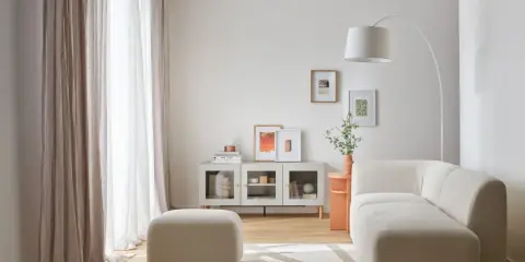 Jasny, długi wąski pokój w bloku z wygodną sofą, pufą i nowoczesną szafką. Zasłony wpuszczają dużo światła.
