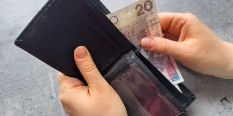 Ręce wkładają banknot 20 zł do portfela. Dowiedz się, komu przysługuje zasiłek dla bezrobotnych.