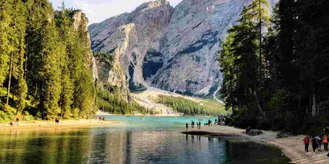 Sentiero da Valdaora al Lago di Braies: acque turchesi, montagne imponenti e pinete rigogliose. Turisti passeggiano sulla riva sabbiosa.