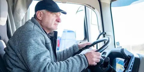 Conductor de camión con gorra y chaqueta gris, con las manos en el volante. Podría ser parte de un listado de cese de actividad de transportistas.