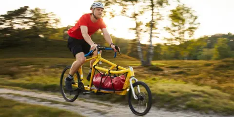Mann fährt mit einem gelben Velo Lab Lastenrad mit roter Tasche durch eine sonnige Landschaft.