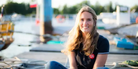 Irina Gracheva, souriante, assise sur un ponton en bois, avec des bateaux et des bouées en arrière-plan.