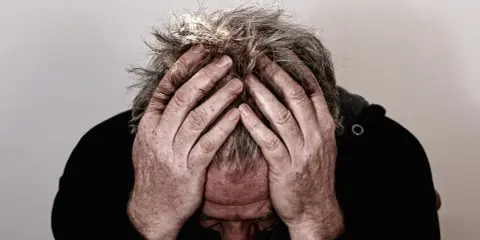Uomo con le mani tra i capelli grigi, testa china, espressione di profonda preoccupazione. Mala tempora currunt sed peiora parantur.