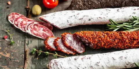 Assortiment de charcuteries, dont une spécialité des Pyrénées enrobée d'épices, sur fond de bois rustique avec tomates et herbes.
