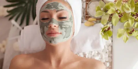 Femme détendue avec un masque fait maison, enveloppée dans une serviette, entourée de fleurs d'orchidée et de bougies.