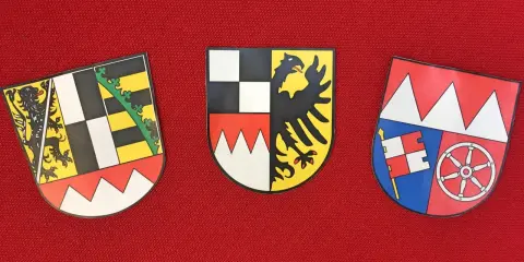 Drei Wappen auf rotem Grund: links ein Löwe, Mitte ein Adler, rechts ein Fähnlein und ein Rad. Sie repräsentieren bayerische Regierungsbezirke.